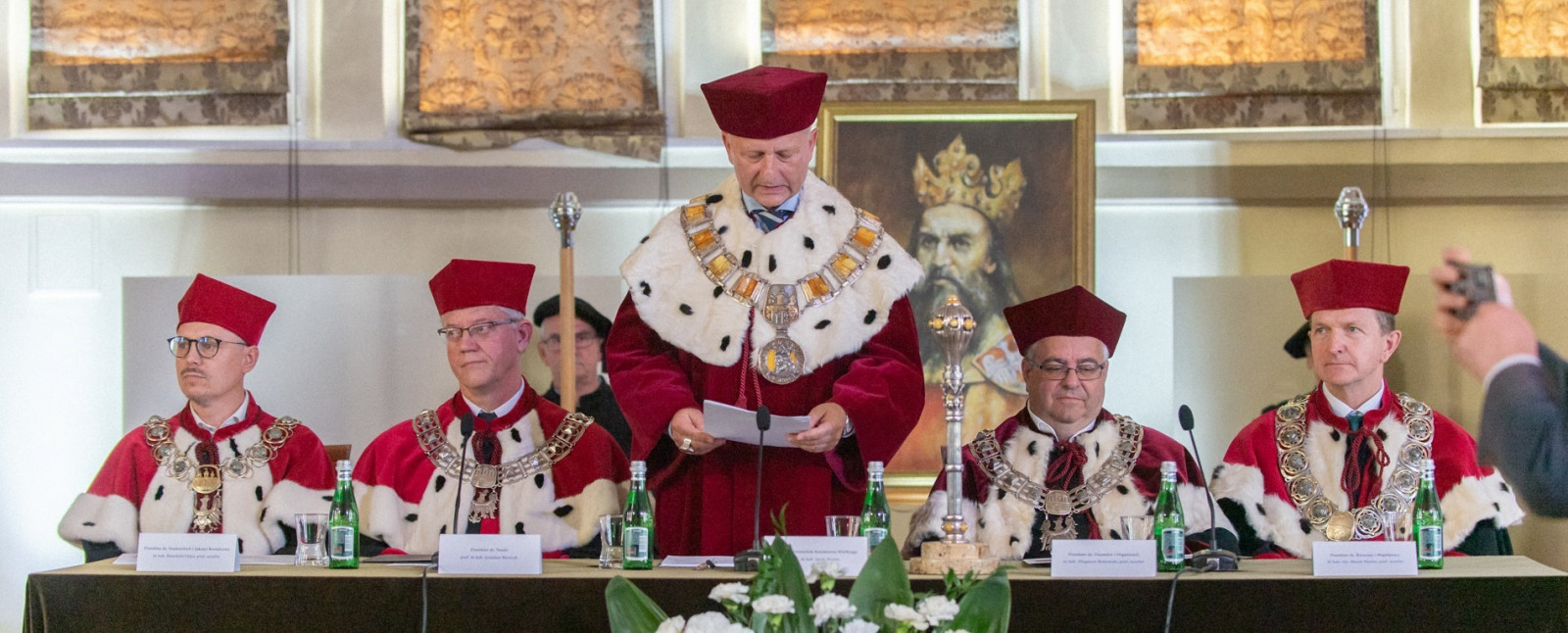 Kolegium retorskie, panowie ubrani w togi rektorskie siedzą przy stole prezydialnym, rektor stoi i przemawia